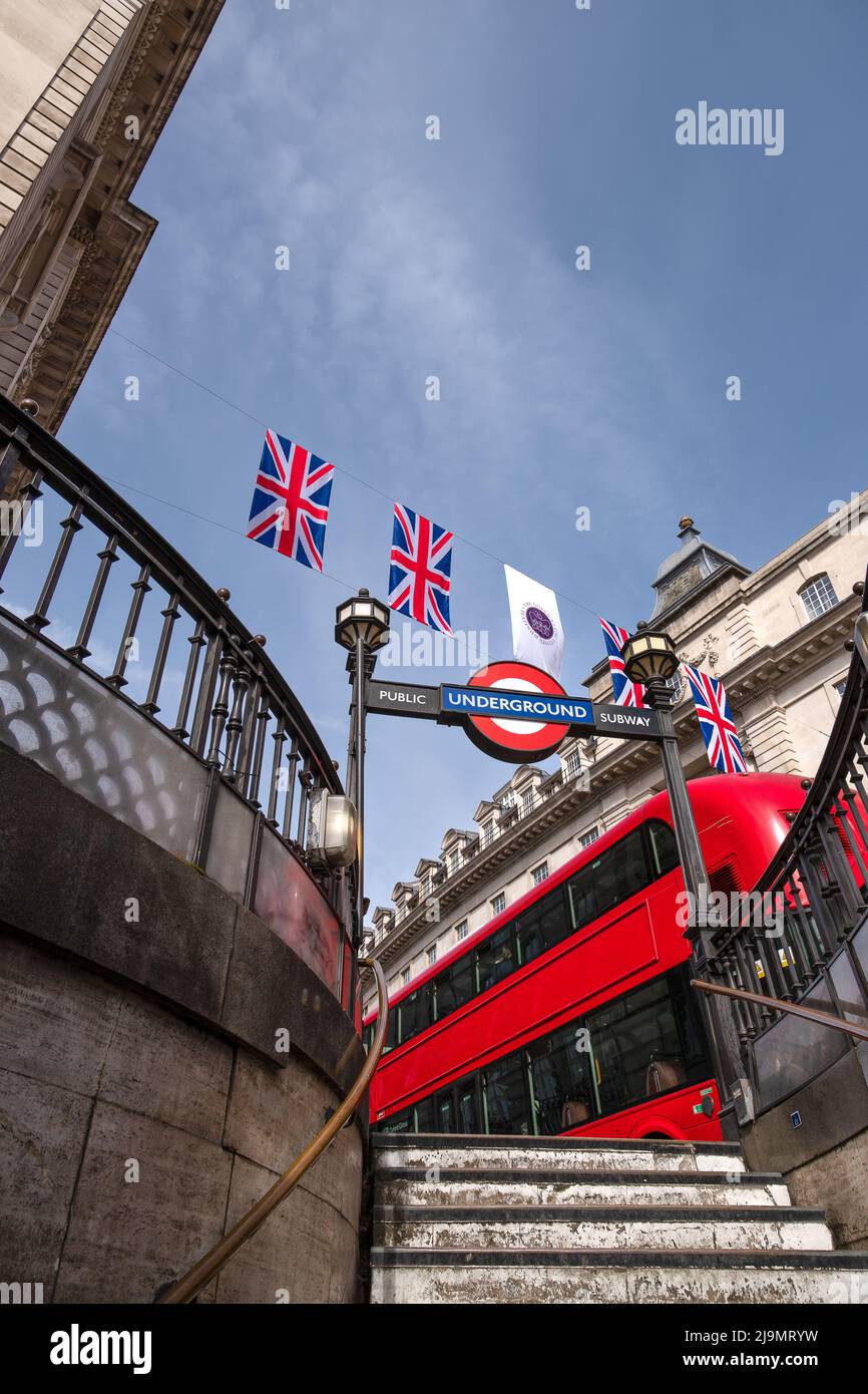 Bunting für das Queen`s Platinum Jubilee über der Piccadilly U-Bahnstation mit einem roten Londoner Bus, der am 21.. Mai 2022 vorbeifährt. Stockfoto